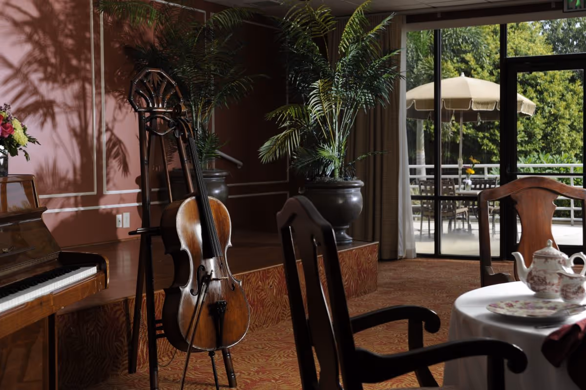 A cozy interior room with a wooden piano and a cello resting on a stand. There are large potted plants near a window that looks out onto an outdoor patio with tables, chairs, and umbrellas. In the foreground, there is a table set with a teapot, cups, and plates, surrounded by wooden chairs.