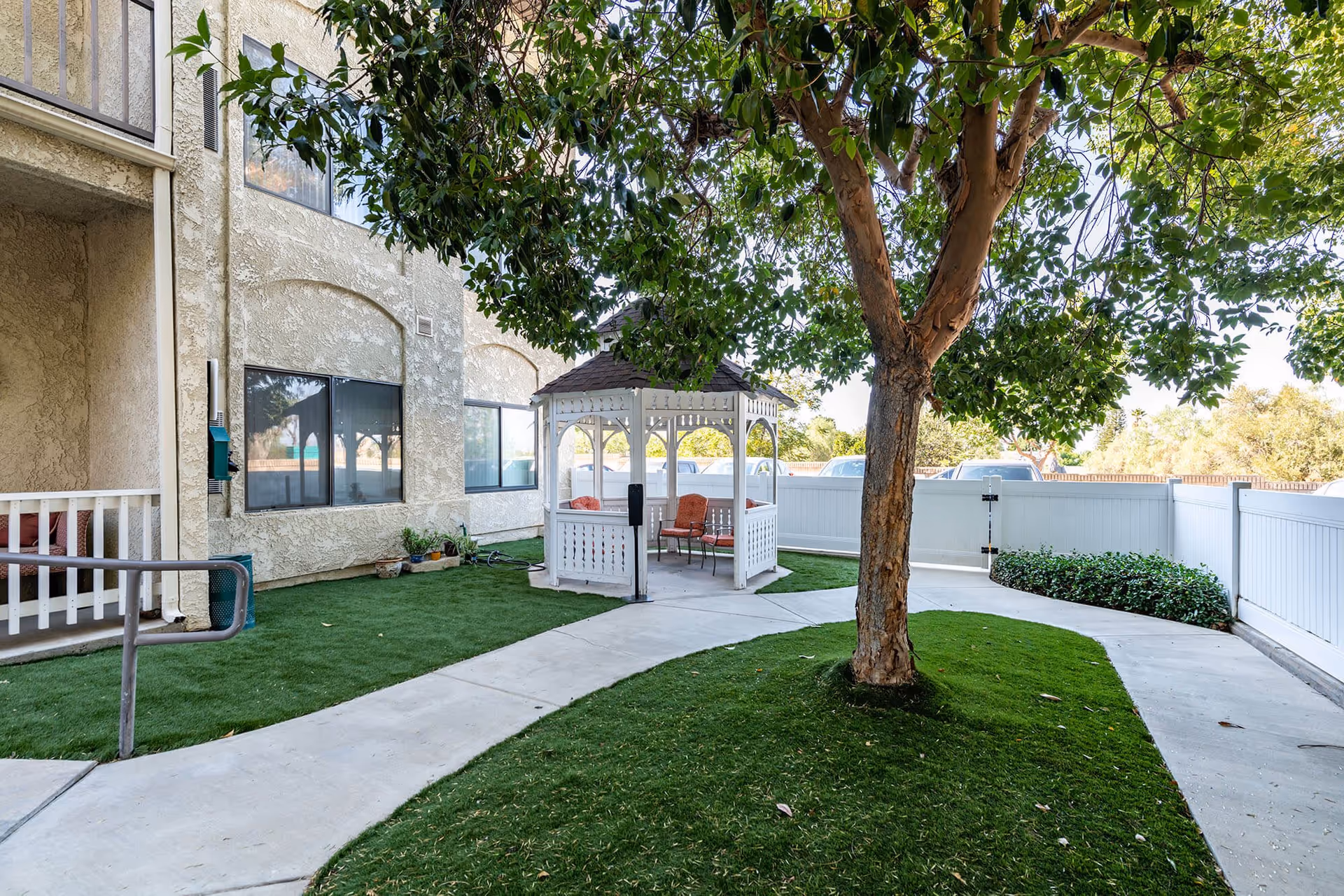 Outdoor courtyard area at Vista Corona Senior Living featuring a large tree, a white gazebo with chairs inside, green grass, a concrete pathway, and a beige building with windows in the background.