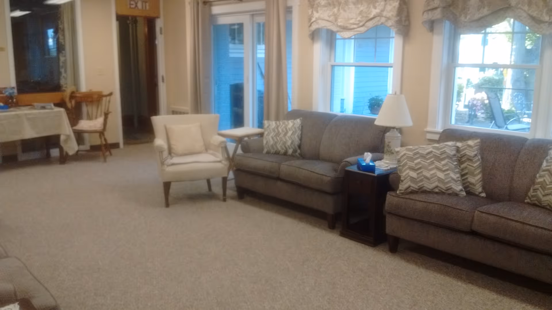 Spacious common room with sofas, an armchair, side tables, lamp and windows.