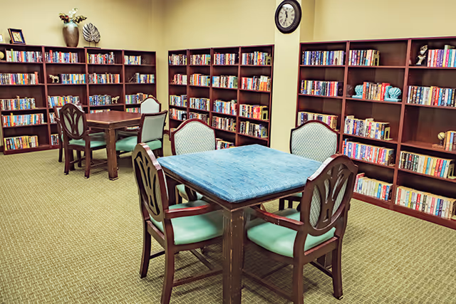 A cozy library room with multiple wooden bookshelves filled with books lining the walls. There are several wooden tables with green cushioned chairs arranged around them. A clock is mounted on the wall above the bookshelves.