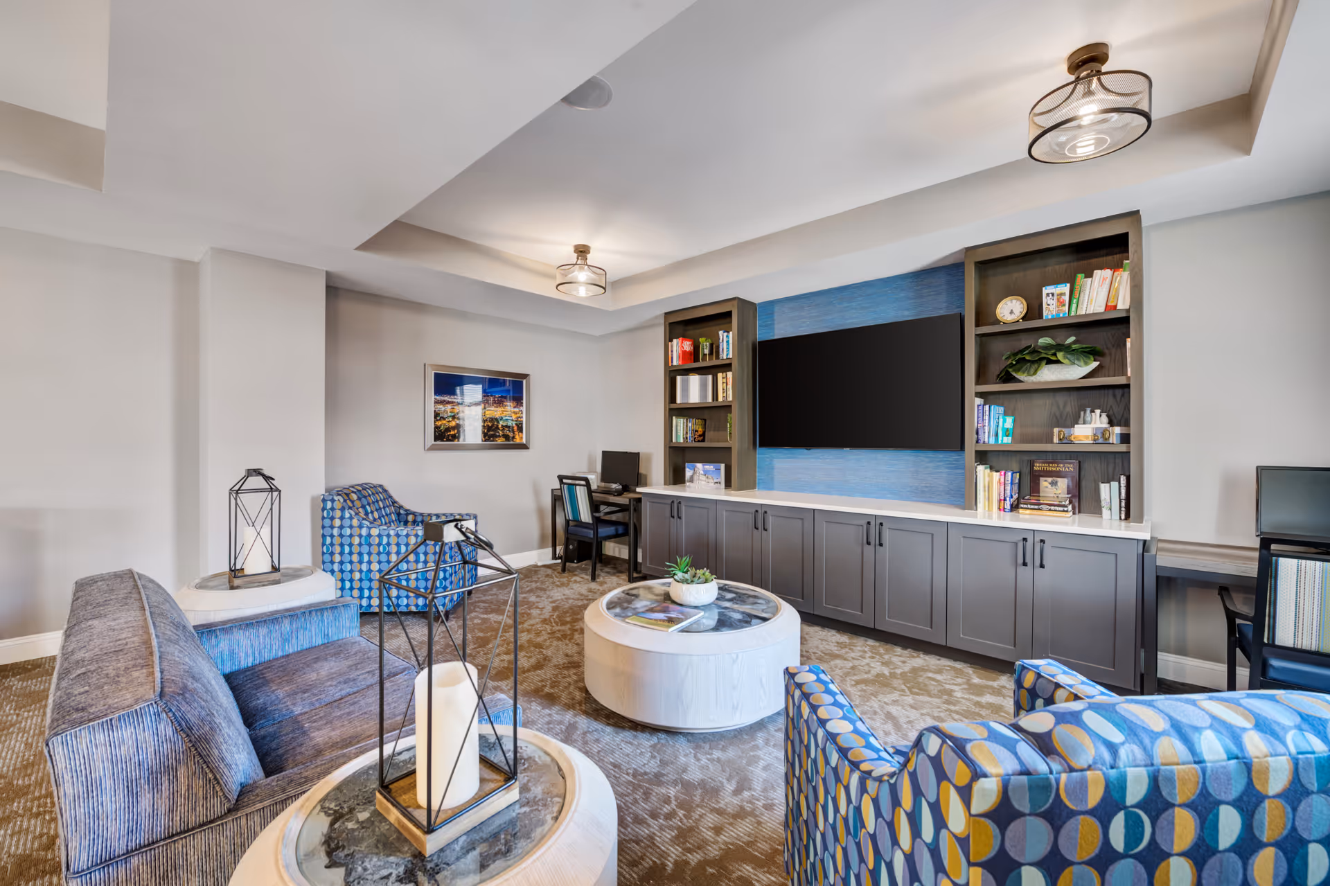A cozy living room area with patterned blue armchairs, a blue sofa, two round white coffee tables with decorative items, a large flat-screen TV mounted on a blue accent wall, and built-in shelves filled with books and plants. There are two desks with chairs and computers on either side of the TV unit. The room has beige carpet and modern ceiling lights.