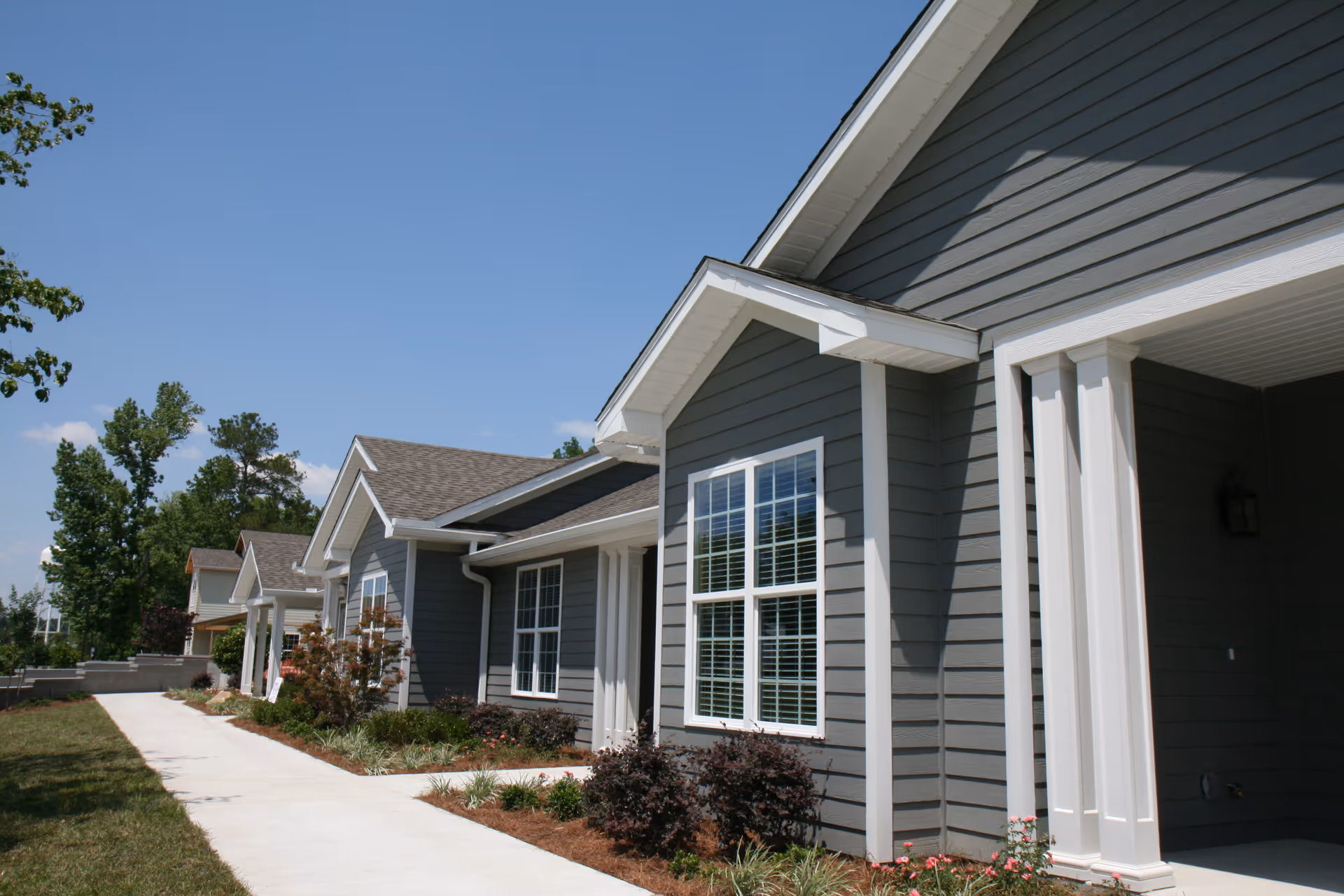 Front exterior of single-story gray residential buildings with white trim, a sidewalk, and landscaped beds under a clear blue sky.
