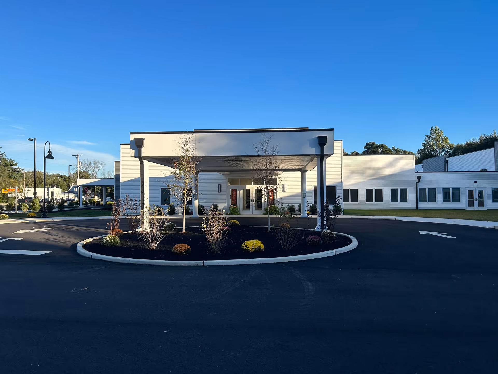 Front exterior view of New Standard Senior Living at Hammonton building with a covered entrance, landscaped roundabout with small trees and shrubs, clear blue sky, and paved driveway.