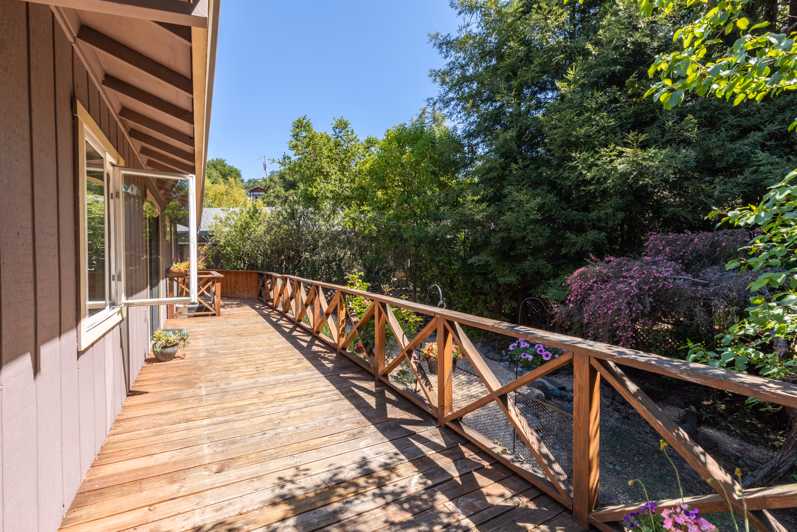 Sunlit wooden deck with a crisscross railing alongside a house, overlooking a lush garden with flowering shrubs.