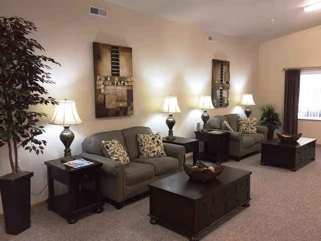 A well-lit living room with two gray sofas each adorned with patterned throw pillows. Between the sofas are dark wooden side tables with lamps on them. Two abstract paintings hang on the beige wall above the sofas. In front of each sofa is a dark wooden coffee table with decorative bowls. A large potted plant is placed on the left side near the wall, and a window with dark curtains is on the right side of the room.