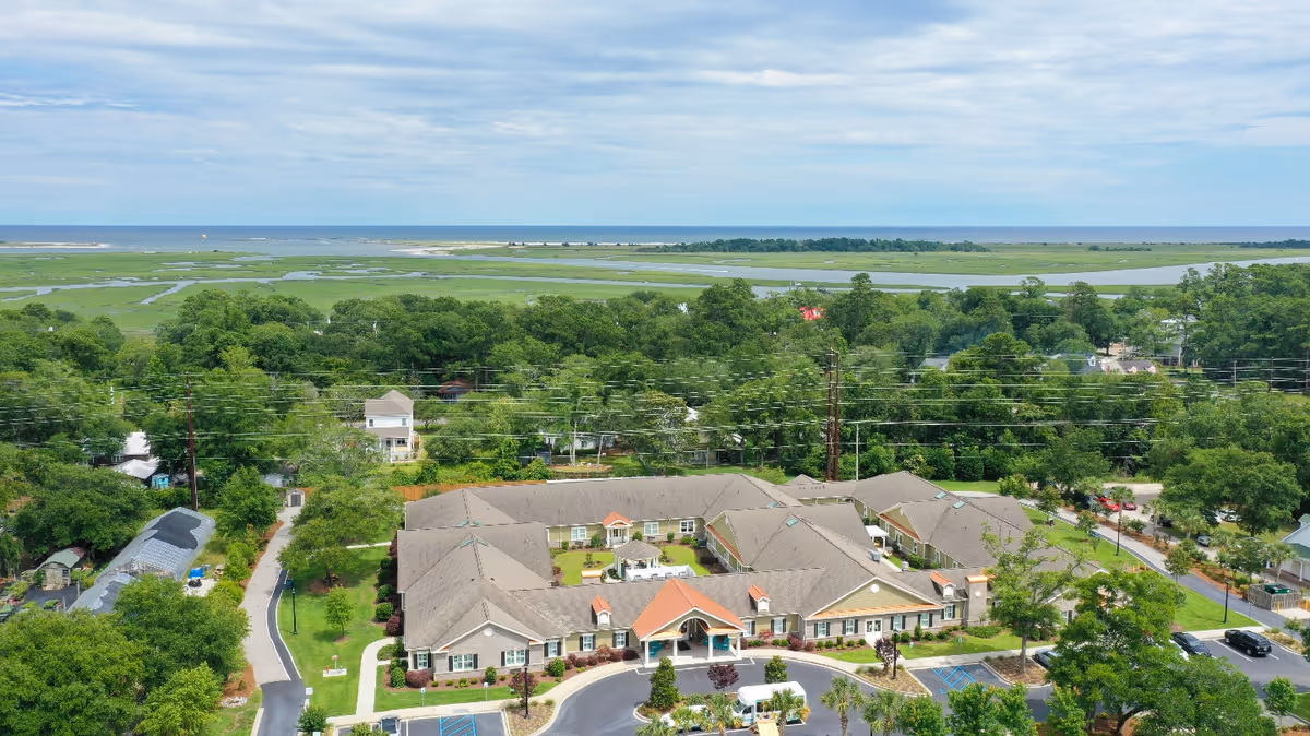 Aerial view of Inlet Coastal Resort Assisted Living and Memory Care facility surrounded by greenery, with a marshland and ocean visible in the background under a partly cloudy sky.