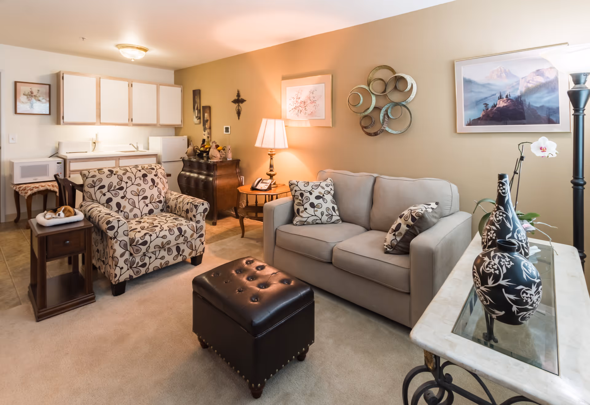 A cozy living room area with a beige sofa adorned with patterned pillows, a matching armchair, and a black leather ottoman. The room features a small kitchenette with white cabinets, a microwave, and a mini fridge. Decorative items include framed artwork, a table lamp, a cross on the wall, and vases on a glass-top table. The space is warmly lit and carpeted.