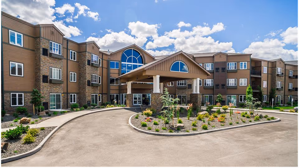 Front exterior view of a large, modern senior living facility building with multiple windows, stone and wood siding, a covered entrance, landscaped garden beds, and a curved driveway under a partly cloudy blue sky.