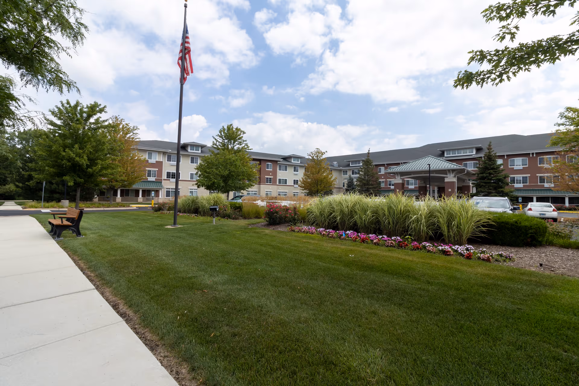 Exterior view of StoryPoint Romeoville senior living facility showing a well-maintained lawn with flower beds, an American flag on a flagpole, benches along a sidewalk, and a multi-story building in the background under a partly cloudy sky.