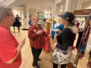 A group of elderly women socializing indoors near a clothing display with mannequins dressed in colorful outfits. The setting appears to be a common area with columns and a piano in the background.