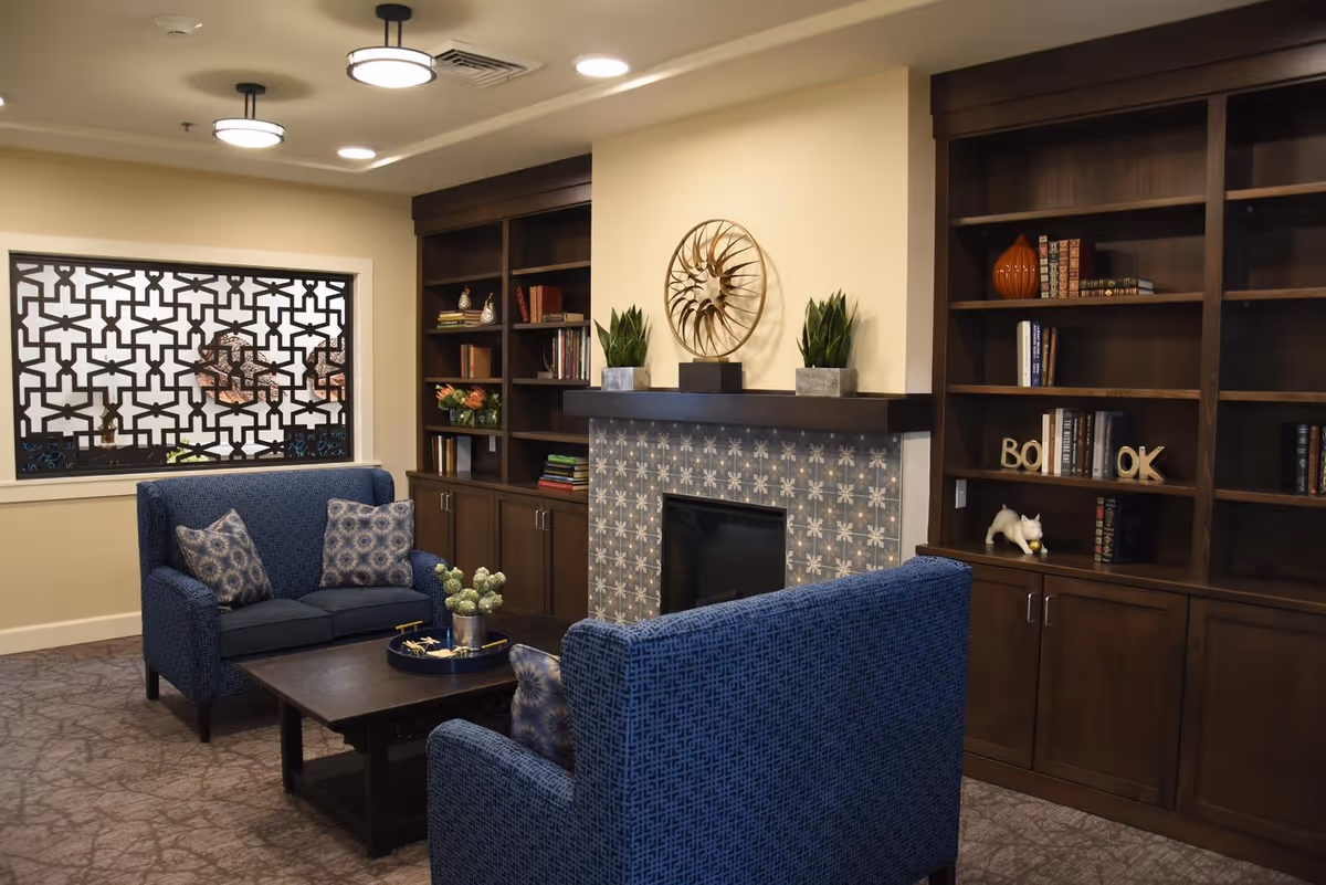 A cozy living room area with two blue upholstered armchairs facing a dark wooden coffee table. Behind the chairs is a fireplace with decorative tile surround and a modern metal wall sculpture above it. Flanking the fireplace are built-in dark wooden bookshelves filled with books and decorative items. A window with a geometric patterned screen is on the left wall. The room has warm lighting from ceiling fixtures and a carpeted floor.