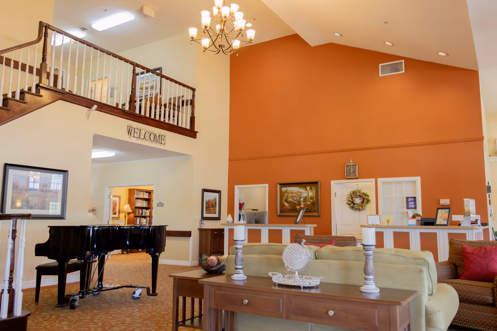 Spacious assisted living facility common area with a grand piano, beige and brown furniture, a wooden table with decorative items, a chandelier, and a welcome sign above a staircase with wooden railing.
