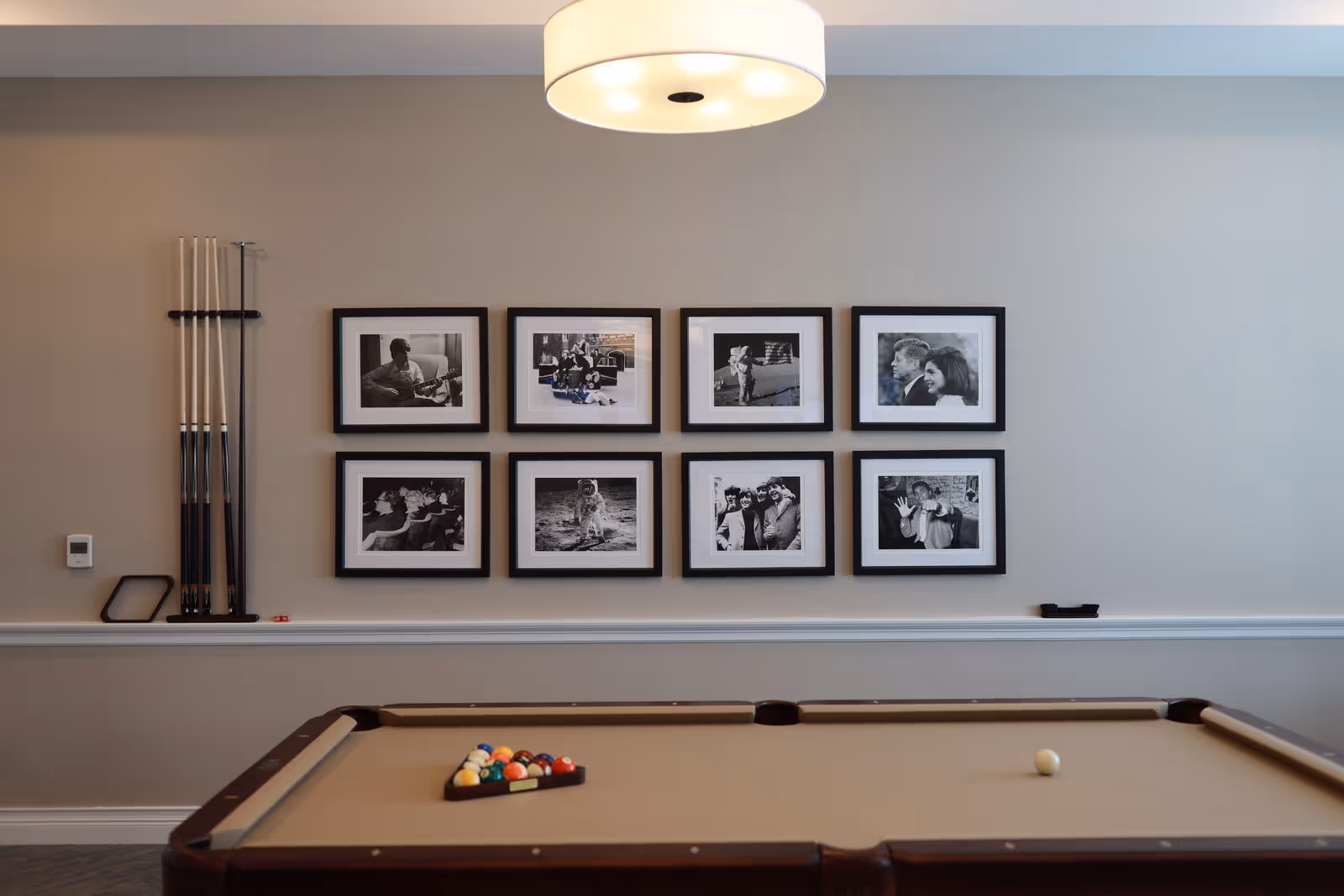 A billiards room with a pool table, cue rack, and a wall of eight framed black-and-white photos under a ceiling light.