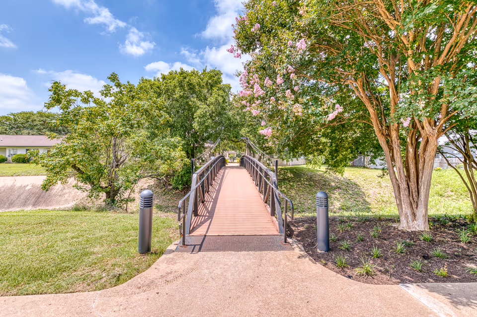 A small pedestrian bridge with metal railings crosses a grassy area with trees and flowering plants on either side under a partly cloudy blue sky.