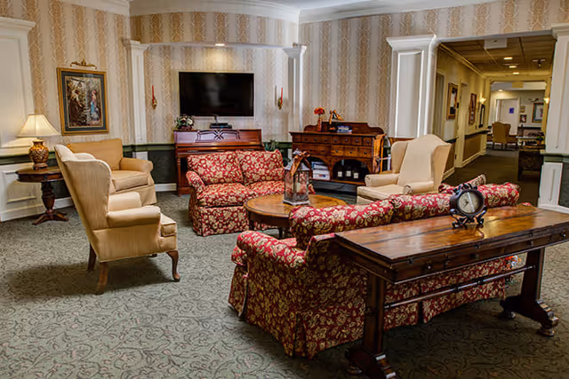 A cozy living room area with patterned wallpaper and carpet. The room features two beige armchairs, two red floral-patterned sofas, a wooden coffee table, a wooden console table with a clock, a piano, a flat-screen TV mounted on the wall, and a wooden sideboard with decorative items. A lamp and framed artwork are visible on the left side, and a hallway extends to the right.
