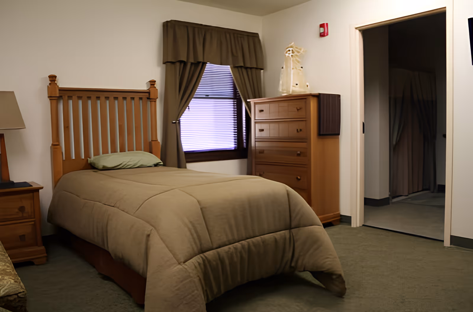 A simple bedroom with a single wooden bed covered with a beige comforter and a green pillow. Next to the bed is a wooden nightstand with a lamp. There is a window with brown curtains and blinds, and a wooden dresser with a decorative item on top. The room has green carpet and white walls, with an open doorway leading to another room.