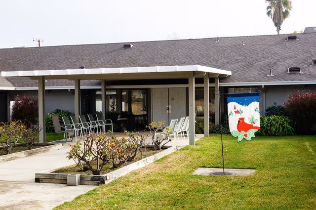 Outdoor view of a single-story assisted living facility building with a covered patio area featuring several chairs arranged in rows. There are trimmed bushes along the walkway and a decorative flag with a red cardinal and winter scene on a pole in the grassy yard.