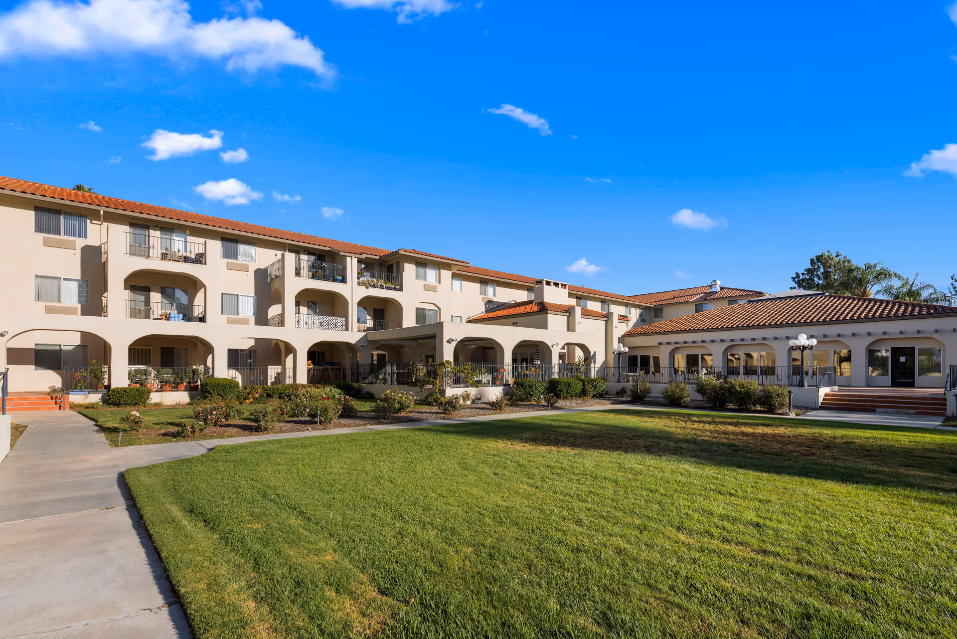 Exterior view of a senior living community building with beige walls and red-tiled roofs under a clear blue sky. The building has multiple balconies and arched walkways. In front, there is a well-maintained green lawn and paved walkways.