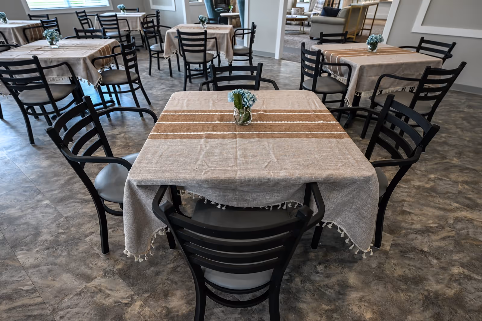 A dining area with multiple square tables covered with beige tablecloths featuring brown stripes. Each table has a small glass vase with blue flowers. Black chairs with gray cushions surround the tables. The floor has a gray marble-like pattern, and there are windows letting in natural light.