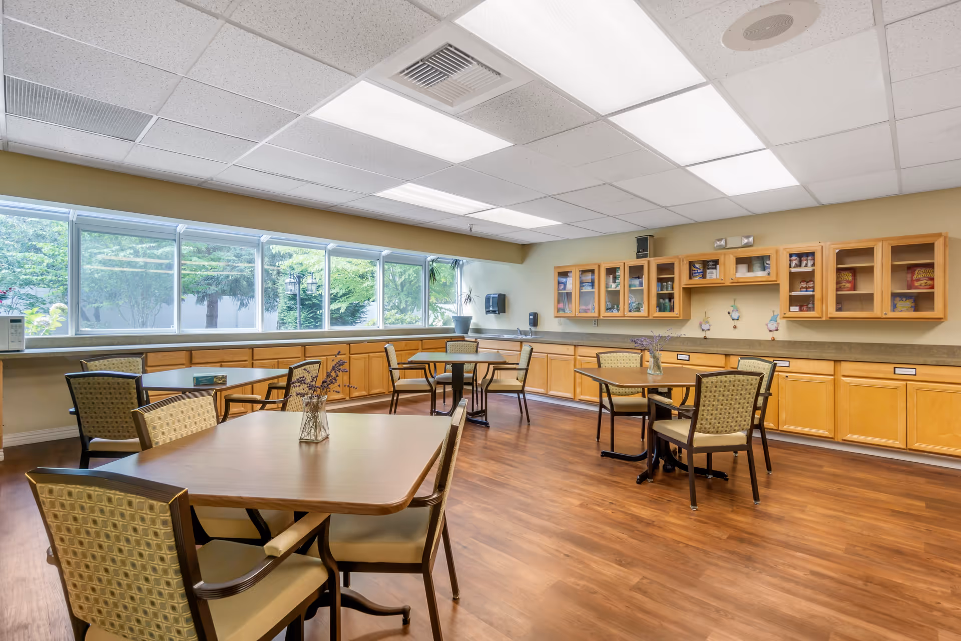 A bright dining room with wooden floors and several tables with chairs arranged around them. The room has large windows along one wall letting in natural light, and wooden cabinets with glass doors on the opposite wall. Small vases with flowers are placed on the tables.