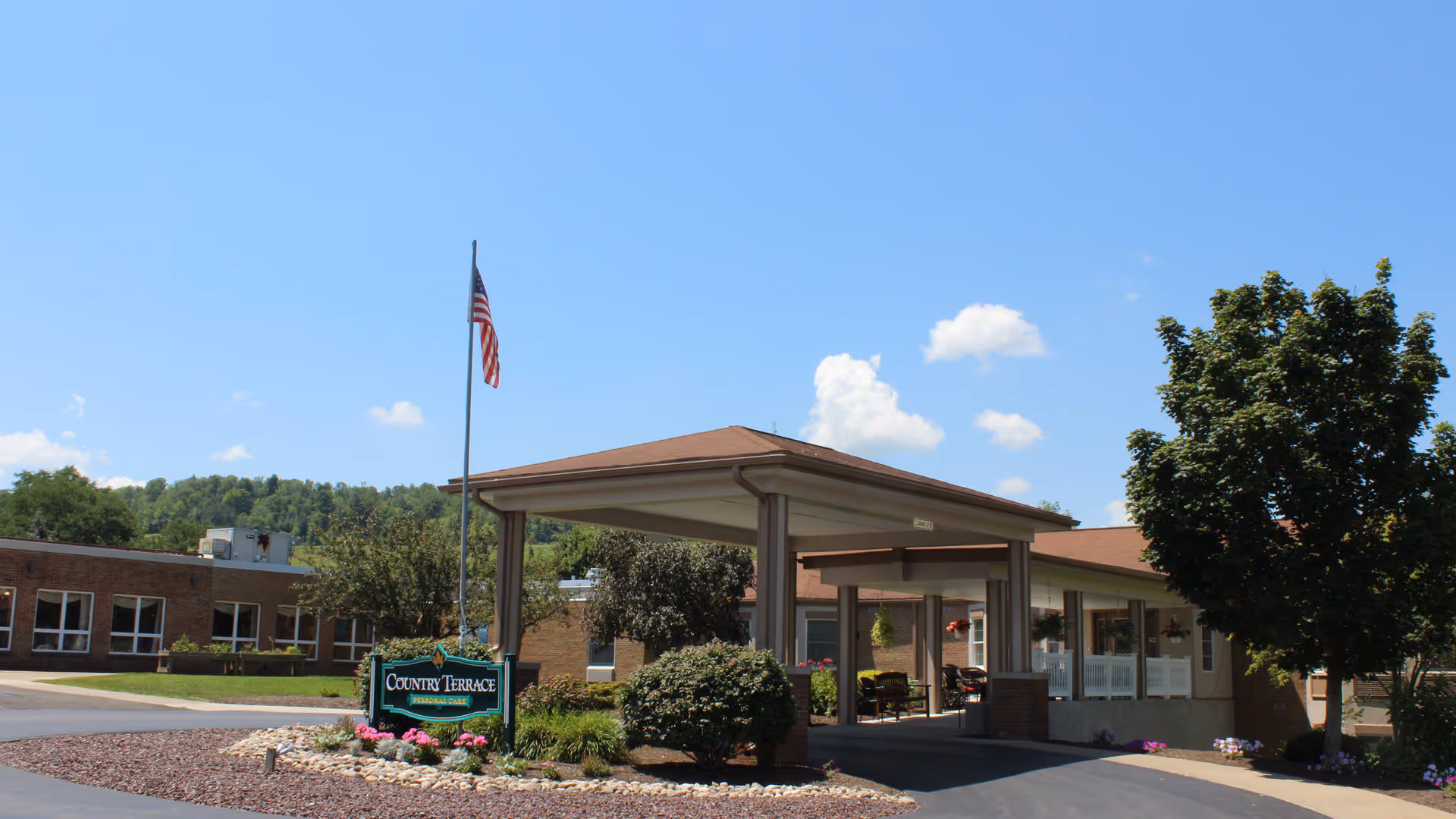 Exterior view of Country Terrace senior living facility entrance with a covered drop-off area, an American flag on a flagpole, landscaping with bushes and flowers, and a clear blue sky with some clouds.