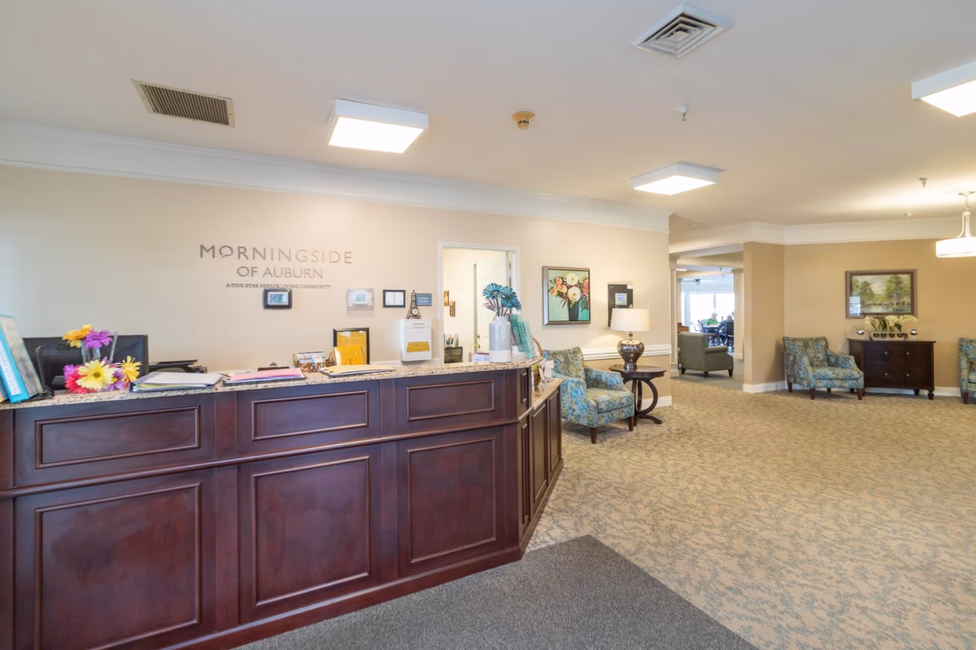 Reception area of Morningside of Auburn senior living community with a dark wood front desk, floral decorations, comfortable seating with patterned armchairs, and soft lighting. The wall behind the desk displays the facility name and slogan.
