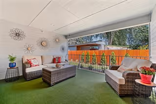 Covered outdoor patio area with artificial grass flooring, featuring two wicker sofas with cushions and pillows, a wicker coffee table, side tables with potted plants, and decorative metal wall art on the white paneled wall. The patio overlooks a fenced backyard with trees and a shed.