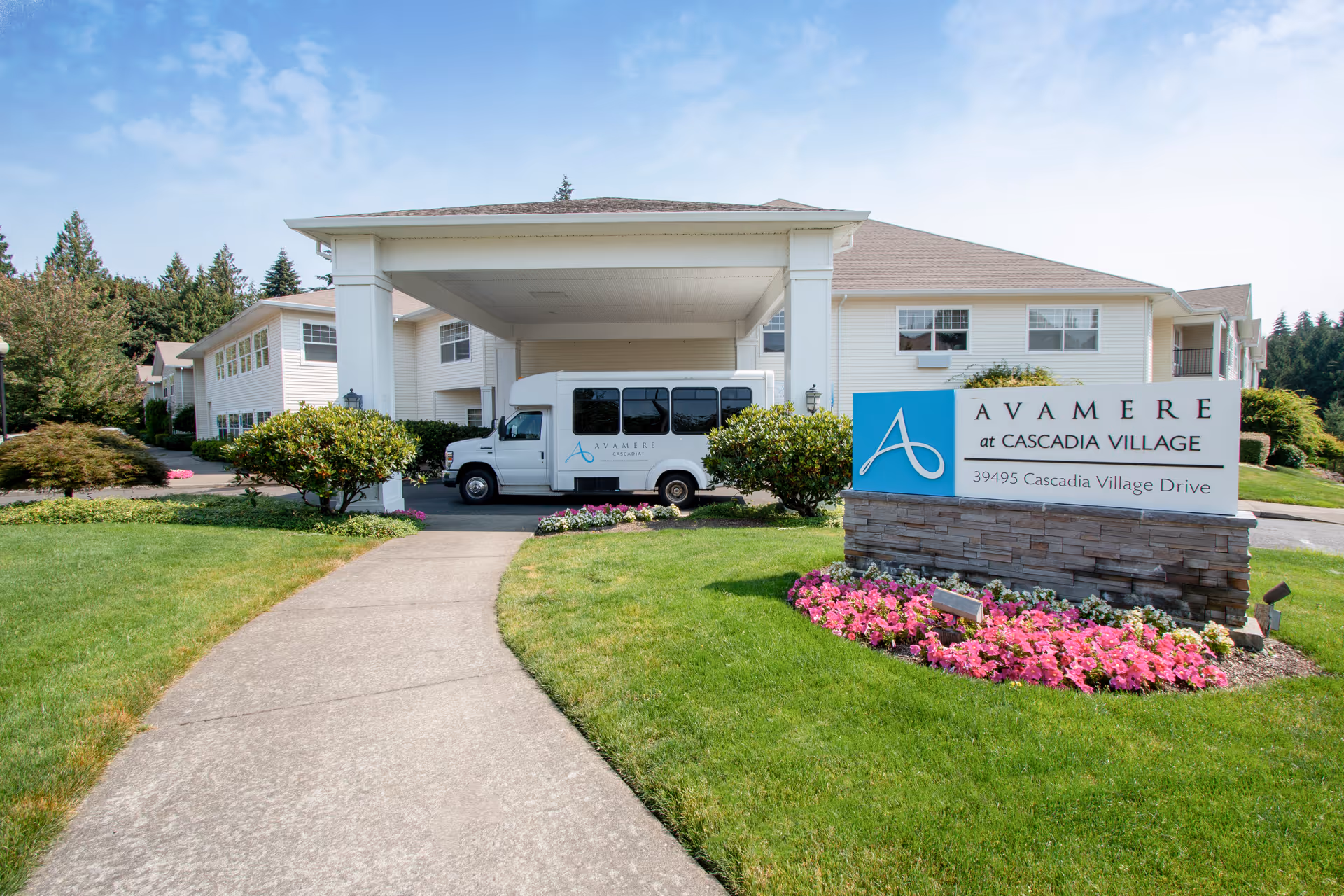 Front exterior view of Avamere at Cascadia Village senior living facility with a covered entrance, a white shuttle van parked under the canopy, well-maintained green lawn, colorful flower beds, and a sign displaying the facility name and address.