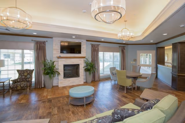 A bright and spacious living room area with wooden flooring, a stone fireplace with a mounted TV above it, two large potted plants on either side of the fireplace, multiple windows with curtains allowing natural light, a round table with four chairs, a green sofa with patterned cushions, an upholstered armchair, and modern ceiling light fixtures.