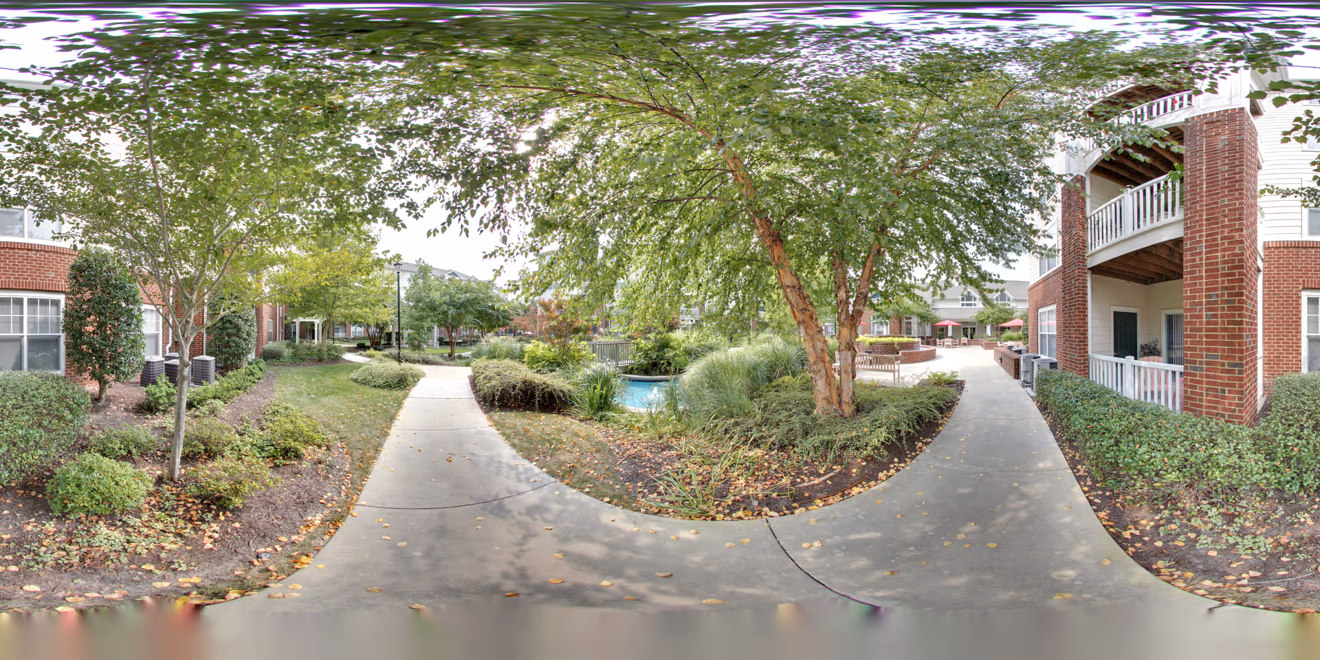 Outdoor pathway in a senior living facility with trees, bushes, and landscaped greenery on both sides. The path splits into two directions, surrounded by red brick buildings with balconies and windows. There is a small water feature and outdoor seating area visible in the background under a cloudy sky.