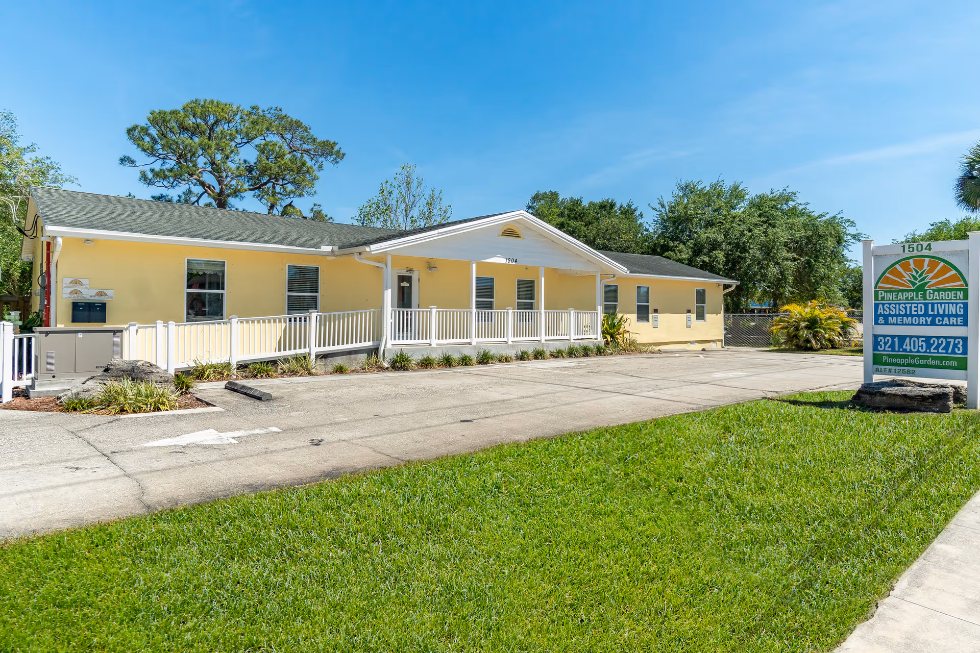 Exterior view of a single-story yellow building with a white railing and a small porch, surrounded by greenery and trees under a clear blue sky. A sign in front reads Pineapple Garden Assisted Living & Memory Care with contact information.