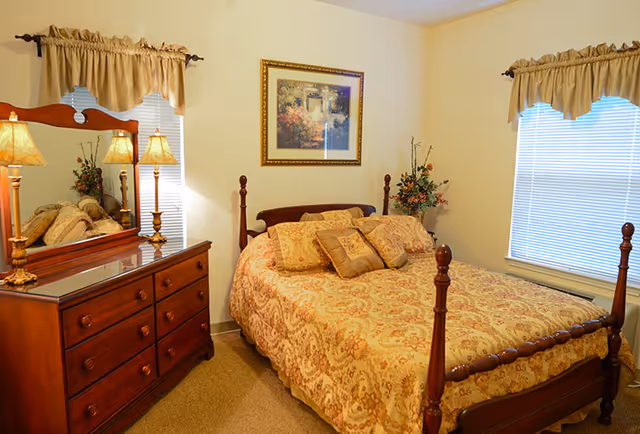 A cozy bedroom with a wooden four-poster bed covered in a patterned beige and gold bedspread with matching pillows. There is a wooden dresser with a large mirror and two table lamps on the left side of the room. Two windows with beige valance curtains allow natural light to enter. A framed floral painting hangs on the wall above the bed, and a floral arrangement is placed in the corner near the window.