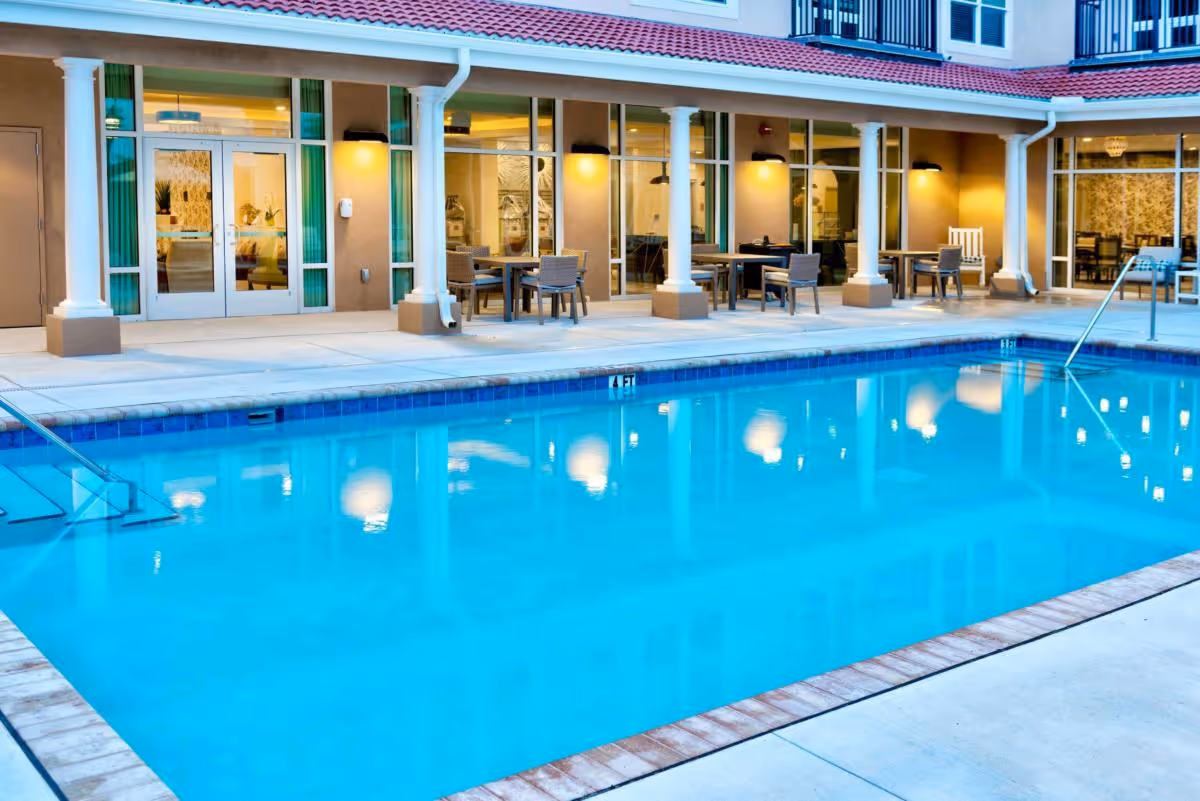 Outdoor swimming pool with a poolside patio, tables and chairs in front of a columned building with glass doors.
