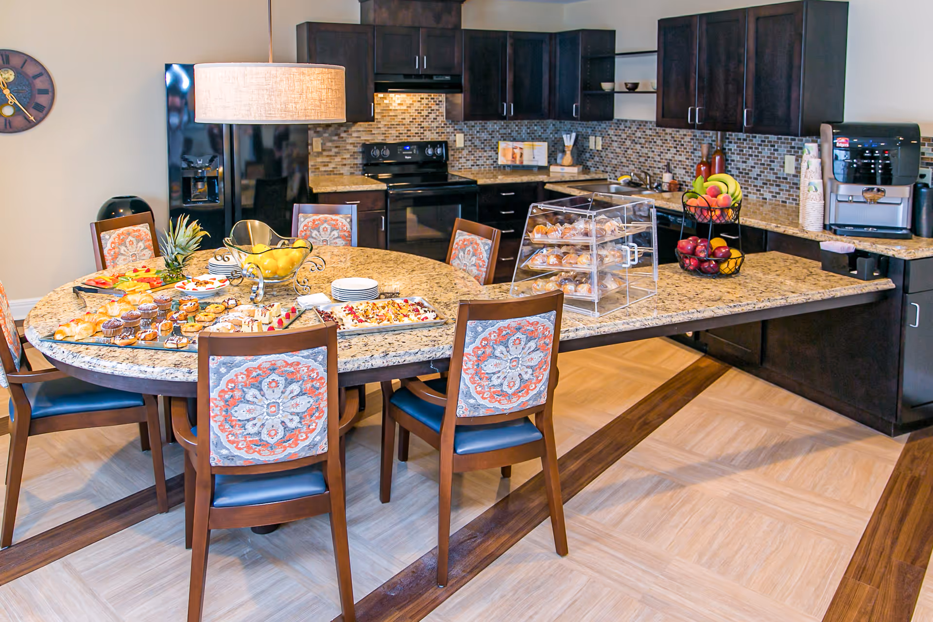 A modern kitchen and dining area with a round granite table surrounded by six wooden chairs with patterned cushions. The table is set with various pastries, fruits, and plates. The kitchen features dark wood cabinets, a black refrigerator, stove, and a coffee machine on the counter. The backsplash is made of small mosaic tiles, and a large pendant light hangs above the table.