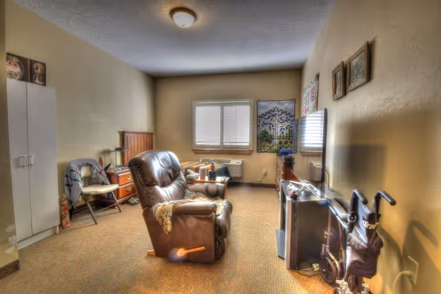 A cozy living room in a senior living facility with a brown leather recliner chair, a small wooden side table with a lamp, a folding chair, a white cabinet, a window with blinds, a TV mounted on the wall, and various framed pictures and decorations on the walls. There is also a walker with a seat and a storage pouch near the TV.