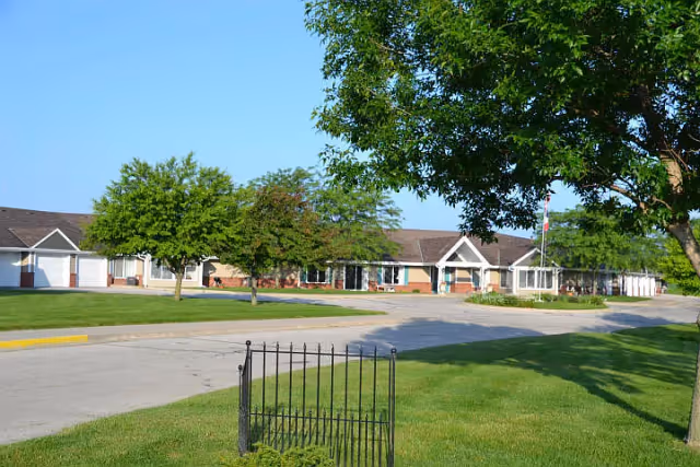Exterior view of Regency Retirement Residence showing a single-story building with a brick and siding facade, surrounded by green lawns, trees, and a paved driveway under a clear blue sky.