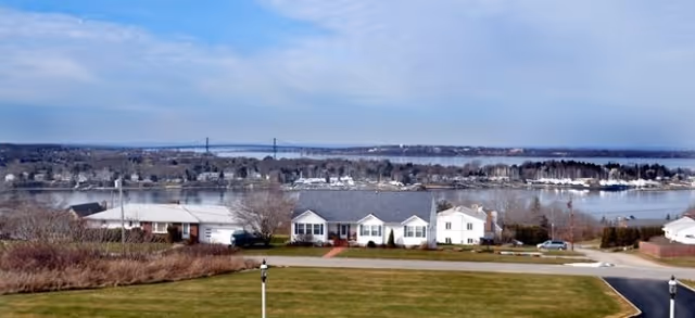 View across a manicured lawn to houses on the shore, a wide bay with boats and a distant bridge under a blue sky.