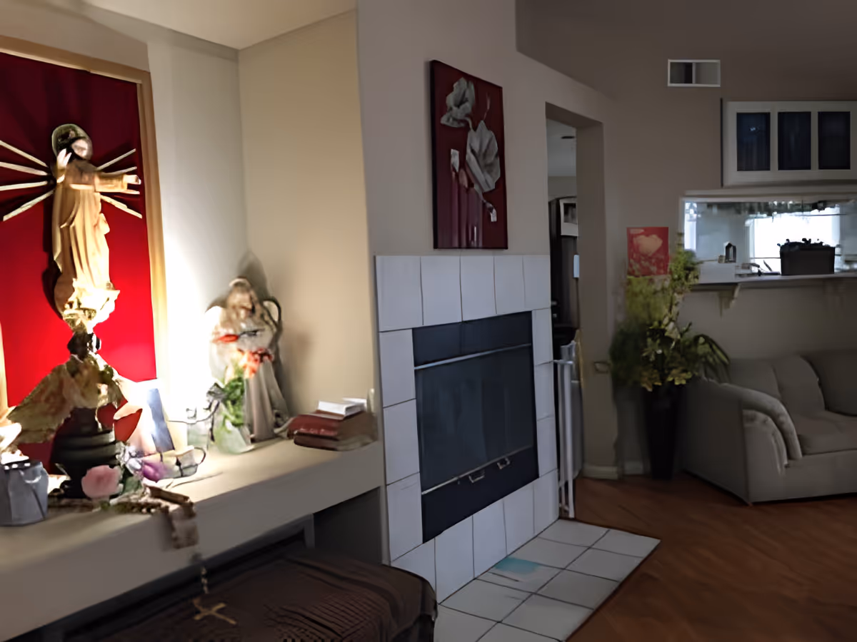 Interior view of a living room area in a care home featuring a white tiled fireplace with a decorative painting above it. To the left, there is a religious statue with a red backdrop and other small decorative items on a shelf. To the right, there is a gray couch and a vase with flowers near a pass-through window to the kitchen.
