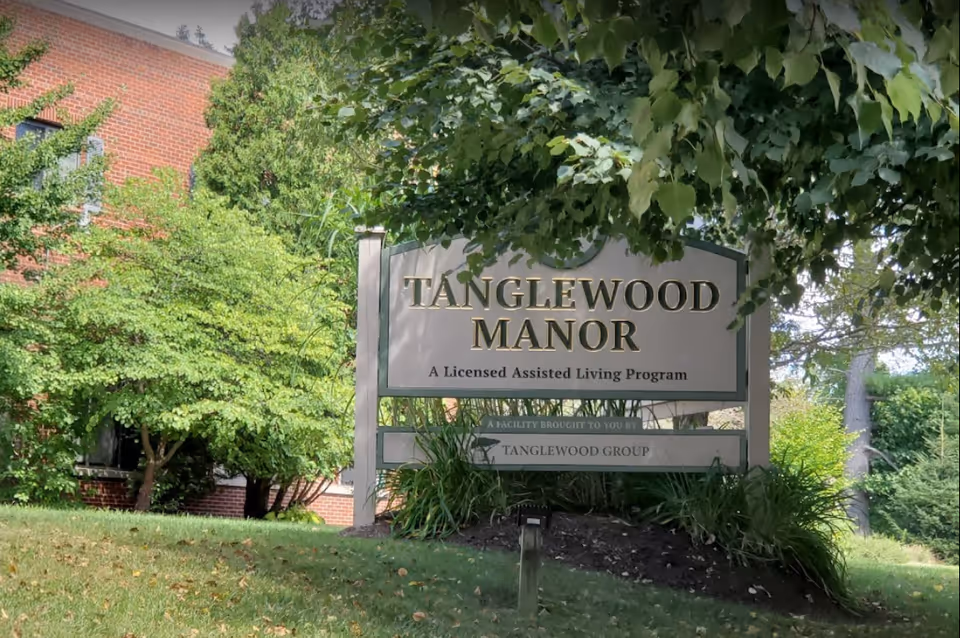 Outdoor view of a sign for Tanglewood Manor, a licensed assisted living program, surrounded by green trees and grass with part of a brick building visible in the background.