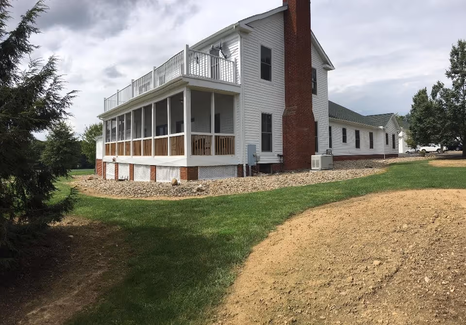 Exterior view of a white two-story building with a large brick chimney, a screened porch with wooden railings, and a balcony on the upper floor. The building is surrounded by green grass, trees, and a patch of bare soil under a cloudy sky.