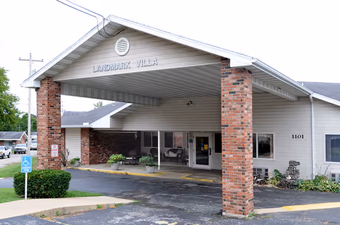 Exterior front entrance of Landmark Villa senior living facility with a covered drop-off area supported by brick columns, a wheelchair accessible parking sign, and some greenery around the building.