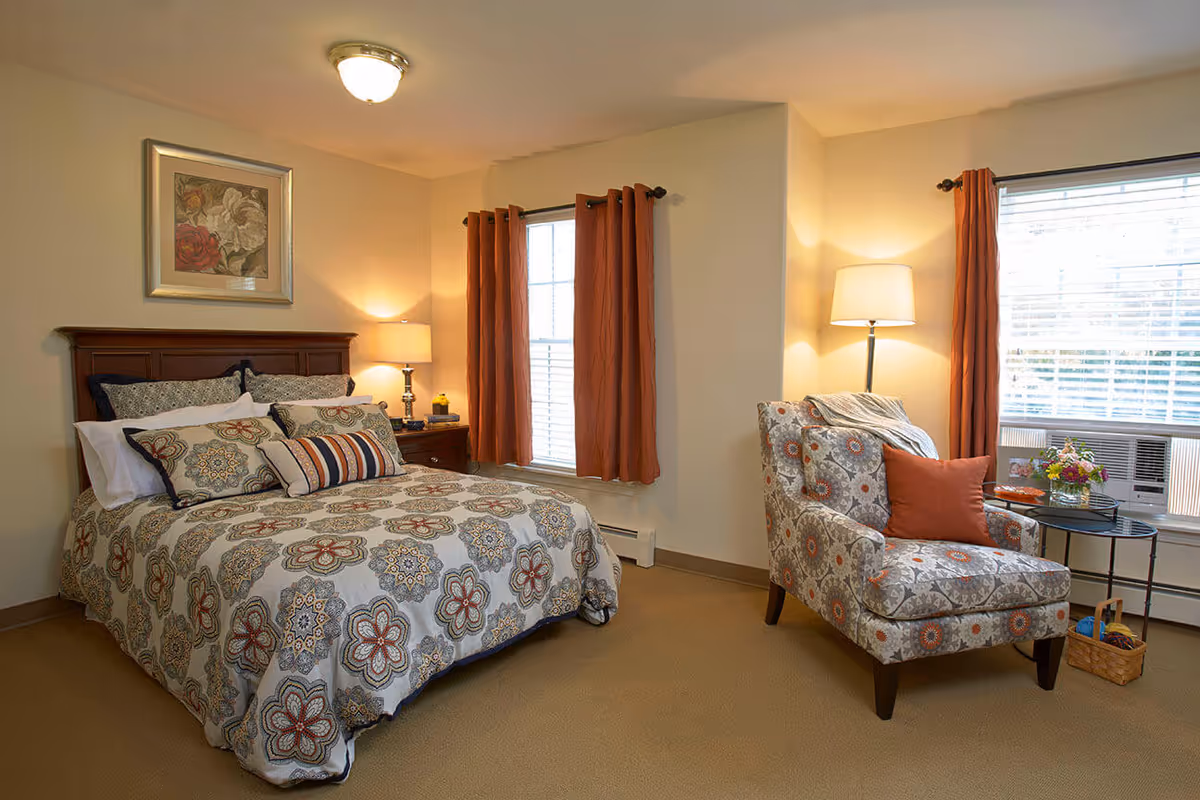 A cozy bedroom with a bed featuring a floral patterned comforter and multiple pillows. There is a wooden headboard and a nightstand with a lamp and decorative items. Two windows with orange curtains allow natural light into the room. A patterned armchair with an orange pillow and a floor lamp are positioned near one window, next to a small round table with flowers and a basket on the floor.