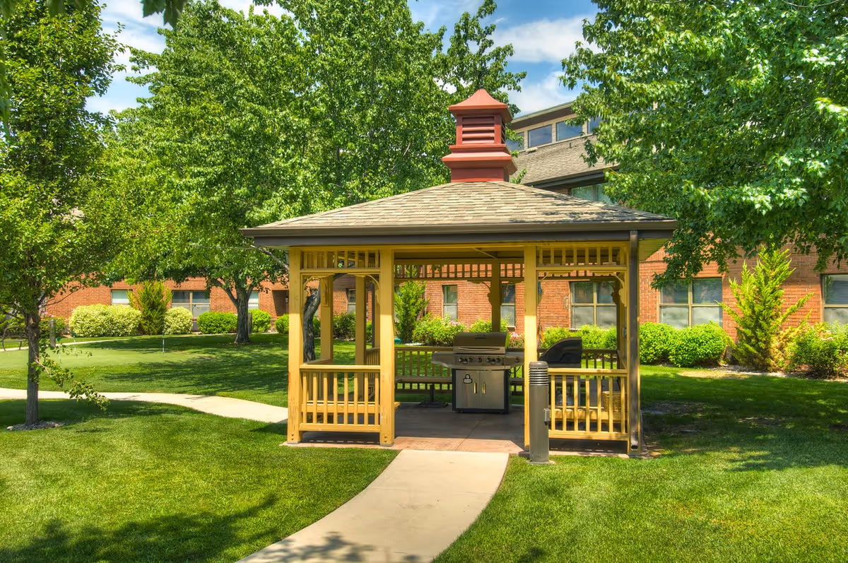 A wooden gazebo with a shingled roof and a red cupola, situated on a well-maintained lawn with a concrete pathway leading to it. Inside the gazebo, there is a stainless steel grill. The background shows a brick building partially obscured by green trees and shrubs under a blue sky with some clouds.