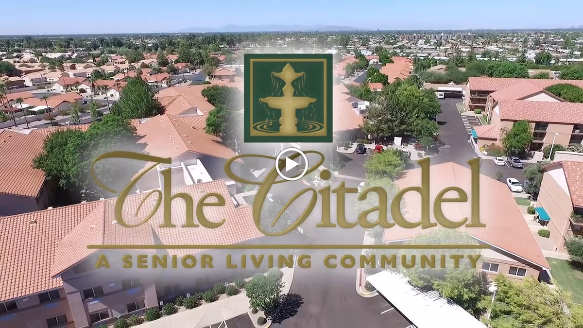 Aerial view of a senior living community with multiple buildings featuring red-tiled roofs, surrounded by trees and parking areas under a clear sky.