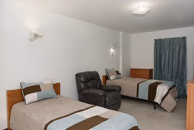 A senior living bedroom with two single beds, each with a beige, brown, and light blue striped bedspread and matching pillows. Between the beds is a dark brown recliner chair. The room has white walls, a wooden dresser, and a window with teal curtains. Two wall-mounted reading lights are above each bed.