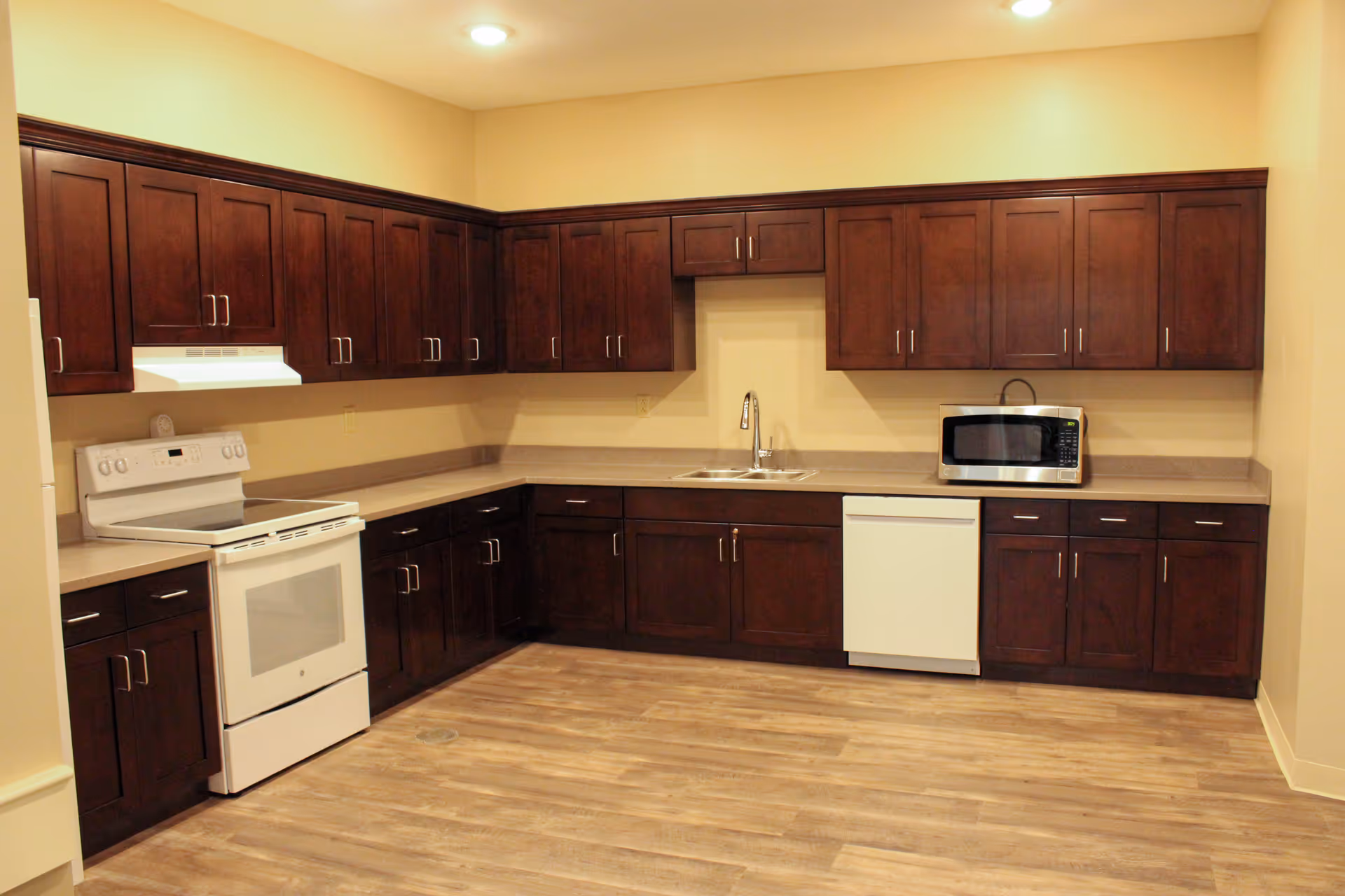 Kitchen with L-shaped dark wood cabinetry, white stove and dishwasher, microwave, sink, and wood-look flooring.