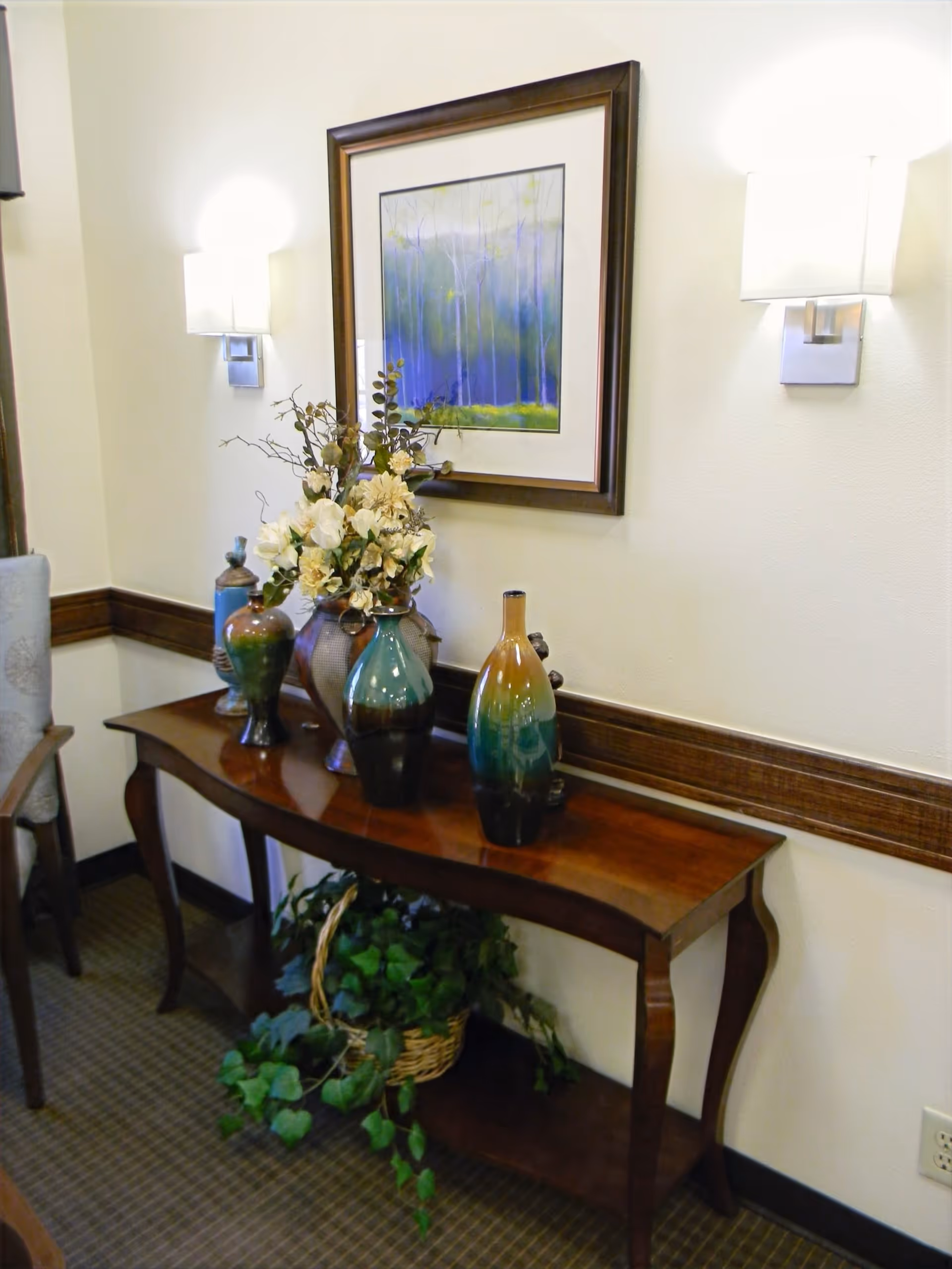 A wooden console table with decorative vases and a floral arrangement on top, placed against a wall with two wall-mounted lights and a framed painting above it. There is a basket with green plants on the lower shelf of the table.