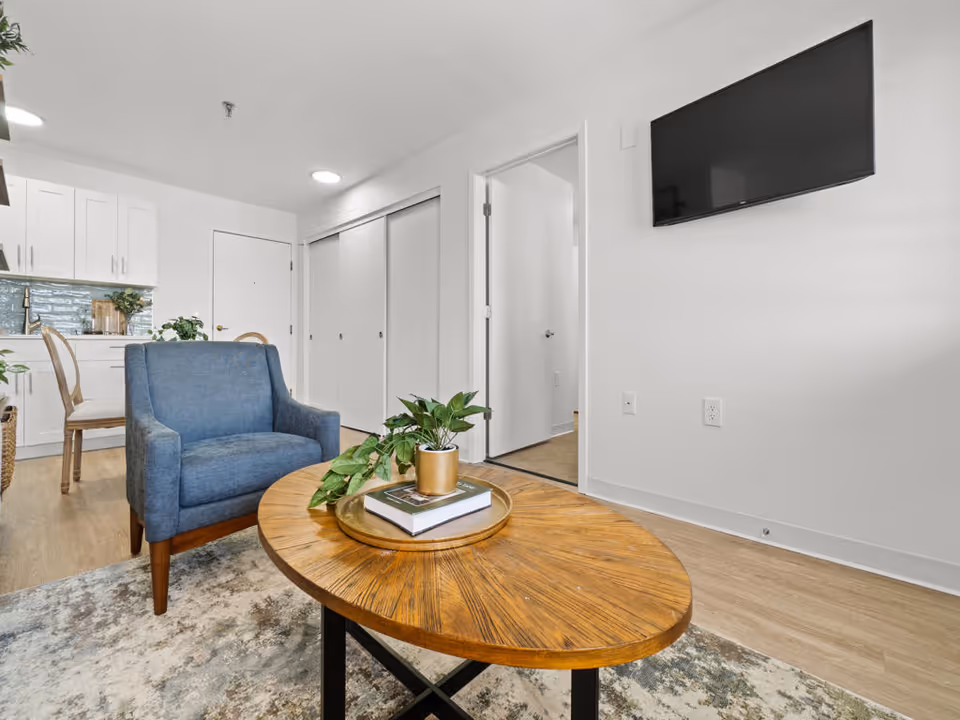A bright and modern living area featuring a blue upholstered armchair, a wooden oval coffee table with a plant and books on a tray, a wall-mounted flat screen TV, and a view into a kitchen with white cabinets and a dining chair. The room has light wood flooring and white walls.