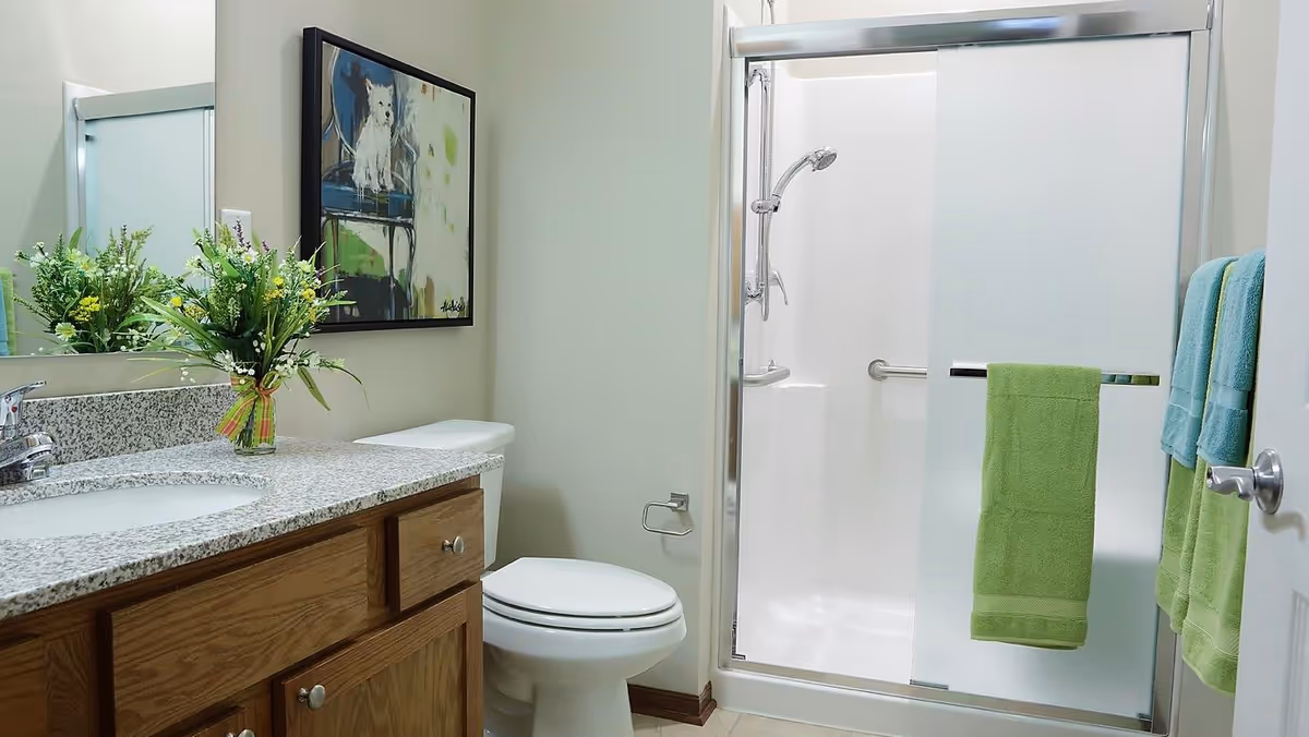 Clean bathroom with a granite-topped vanity, toilet, and sliding frosted glass shower door, accented by green towels and a small floral arrangement.