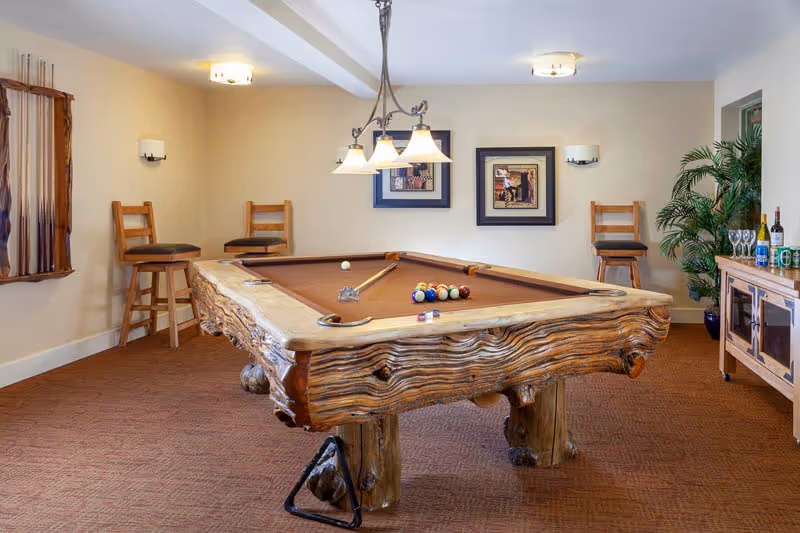 A cozy game room featuring a rustic wooden pool table with billiard balls and cues arranged on it. The room has beige walls, carpeted floor, two framed artworks on the wall, three wooden bar stools, a hanging light fixture above the pool table, a sideboard with bottles and glasses, and a potted plant in the corner.